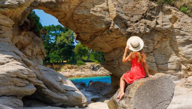 Kırmızı elbiseli kadın sahilde kayanın üzerinde oturuyor. Doğal yaz tatili, tropikal tatil, rahatlatıcı konsept (Brittany, Fransa)