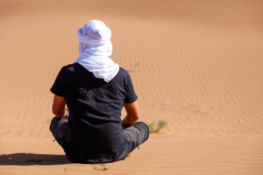 Beyaz türbanlı beyaz bir adam oturmuş çöle bakıyor.