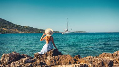 Şapkalı ve beyaz elbiseli bir kız denizdeki yata bakıyor.