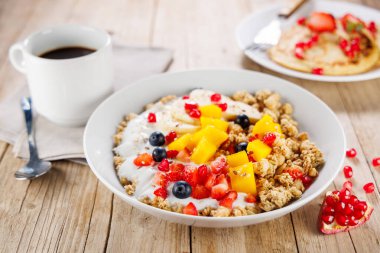 Bir kase granola yoğurt ve taze meyve, ahşap arka planda kahve fincanı.