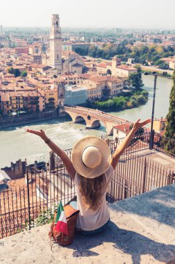 Şapkalı mutlu kadın turist, İtalyan bayrağı ve kolları havada sırt çantasıyla ünlü İtalyan şehir manzarasının panoramik manzarasına bakıyor. Verona.