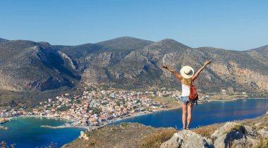 Şapkalı, sırt çantalı ve kolları havada, deniz ve şehrin güzel manzarasının tadını çıkaran genç yürüyüşçü kadın panoramik manzarası. Seyahat yeri, turizm, Yunanistan gezisi