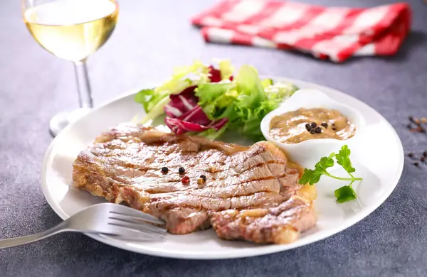 Izgara biftek, biber sosu ve salata, masada bir bardak ve çatal şarap.