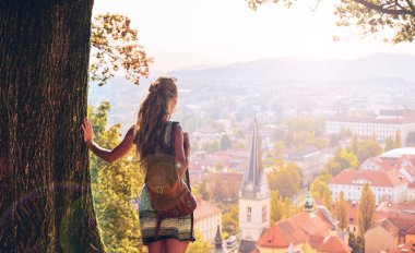 Slovenya 'daki Ljubljana' da panoramik şehir manzarasına bakan bir ağaca karşı duran genç kadın