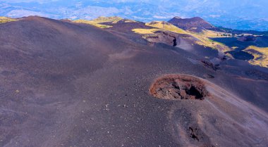 Sicilya 'da açık mavi bir gökyüzünün altındaki yeşil tepeyle çelişen siyah kül manzaralı ve kraterli etna yanardağının hava görüntüsü.
