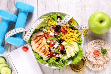 Kızarmış tavuk salatası, avokado, mısır ve feta ile sağlıklı fitness öğle yemeği, halterlerle çevrili, mezura, pirinç keki ve beyaz ahşap arka planda elma