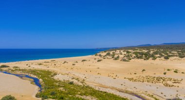 Dağınık çalıları olan geniş kumlu sahil ve açık mavi gökyüzünün altında denize akan küçük bir nehir, yazın vahşi kıyı manzarası.