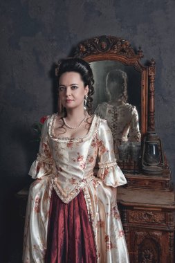 Rokoko tarzı ortaçağ elbisesi giymiş genç ve güzel bir kadın konsol ayna masasının yanında duruyor.