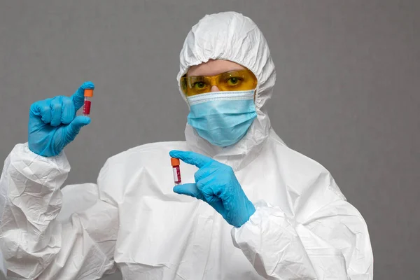 Doctor in protective suit and medical mask with gloves and yellow glasses holding a test tubes with blood and makes an analysis