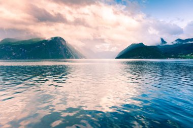 Norveç 'teki fiyort güzelmiş. Berrak mavi su, dağlar ve güneş batarken bulutlu gökyüzü. Yaz manzarası 