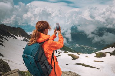 Sırt çantalı genç bir gezgin dağın tepesinde duruyor ve akıllı telefonuyla fotoğraf çekiyor. Dağların yaz manzarası ve bulutlu gökyüzü. Seyahat ve macera konsepti