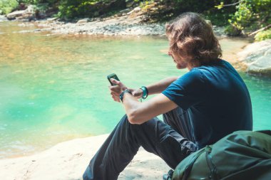 Sırt çantalı genç bir gezgin dağ gölünün kayalıklarında oturuyor ve akıllı telefon kullanıyor. Yaz manzarası. Seyahat ve macera konsepti