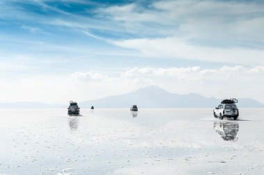 Bolivya 'da Salar de Uyuni' de düz yolda giden off-road arabaları. Gökyüzü ve volkanlar su yüzeyine yansıyor. Meşhur seyahat yeri