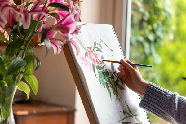 stock image Young girl painting still life with flowers, purple lilies, with watercolor paints on the easel at home