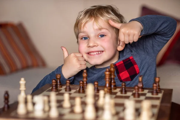 5岁的小孩在大棋盘上玩国际象棋 棋盘放在男孩面前的桌上 他很高兴赢了 竖起大拇指 — 图库照片