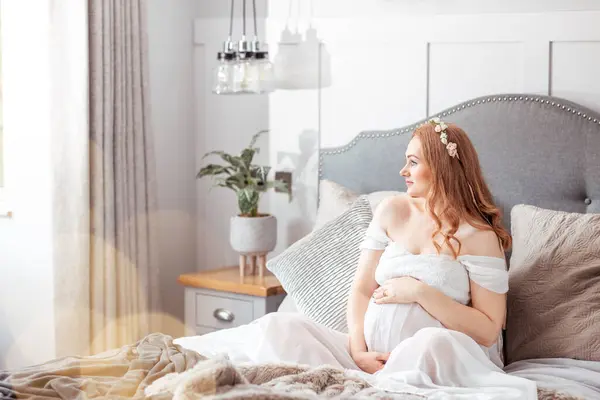 美しい赤毛中年の妊娠中の女性は 自宅でベッドに座っている赤ちゃんを期待しています ストック画像