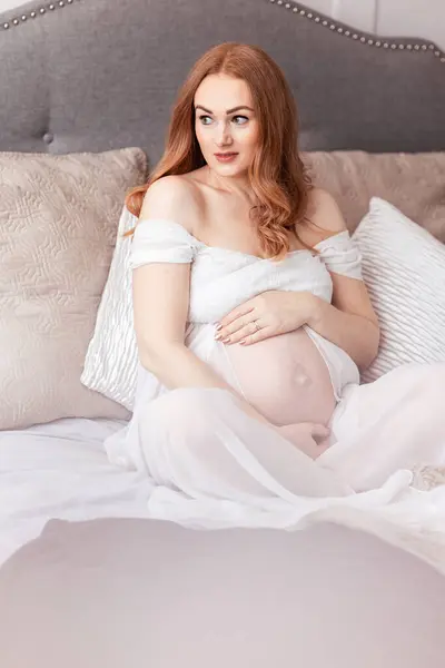 美しい赤毛中年の妊娠中の女性は 自宅でベッドに座っている赤ちゃんを期待しています ロイヤリティフリーのストック画像