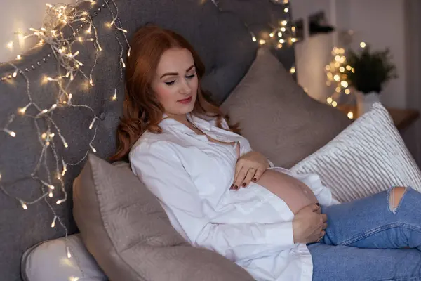 美しい赤毛中年の妊娠中の女性は 赤ちゃんが自宅のベッドに横たわっていることを期待しています ロイヤリティフリーのストック画像