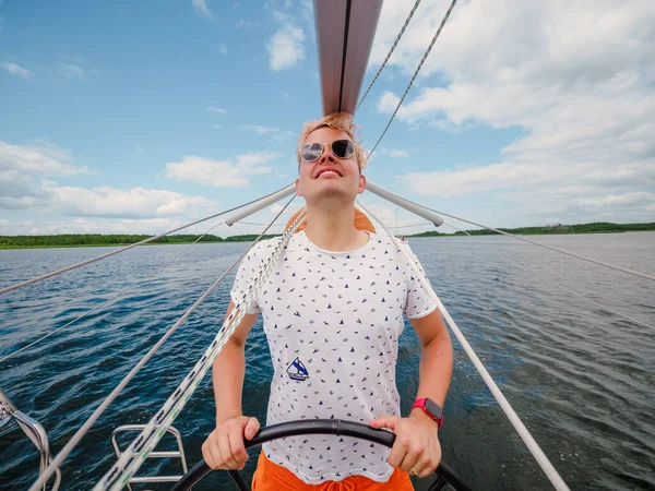 在夏天的航行中 英俊的金发年轻人驾驶帆船或游艇驶过运河 在湖面上挂着折叠的桅杆 — 图库照片