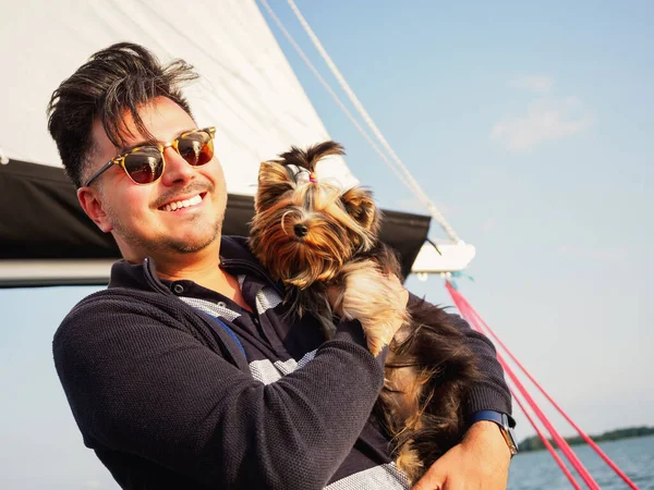 英俊潇洒的人一边度假一边在游艇上抱着他的小狗 约克郡的小狗 一边带着宠物旅行 图库图片
