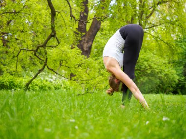 Genç, güzel sarışın kadın güneşli bir yaz gününde parkta yoga yapıyor, Padangusthasana pozunda duruyor.
