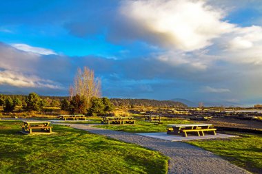 Early spring sunset time, picnic area near the shore , Picnic tables on a green lawn against a mountain range and stormy cloudy sky. This park is located on the Sea Island close to Vancouver Airport clipart