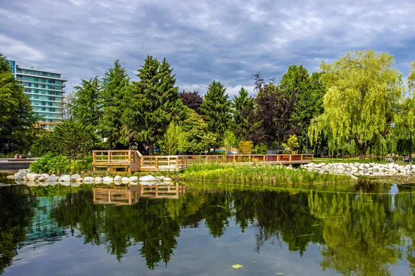 美丽的夏日 城市公园里的一个池塘 一座木头桥 水面上的一条长廊 一棵绿树和一片青草 背景中的建筑物 还有一个戏剧性的云天 — 图库照片