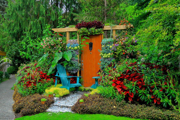 A romantic resting place for two people at Glenbrook Ravine Park in New Westminster City. Two armchairs are located at the decorative door on a green lawn with flower beds among flowering shrubs under the canopy of spreading trees, British Columbia