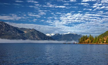 Porteau Cove İl Parkı. Güneşli kış günü, deniz ve dağlar. Körfezdeki mavi su, sis suya çöktü. Arkaplanda karla kaplı bir sıradağ bulutlu güzel gökyüzüne karşı