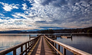  Belcarra Regional Park, Pier on the bay shore against the backdrop of a ridge and cloudy sky, a small village on the opposite bank, floating docks on the sides of the pier