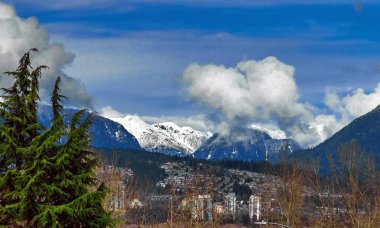 Coquitlam Limanı 'ndaki Mountainside yerleşim bölgesi, Köy ve yüksek binalar karla kaplı dağ sıraları ve mavi bulutlu gökyüzüne karşı.