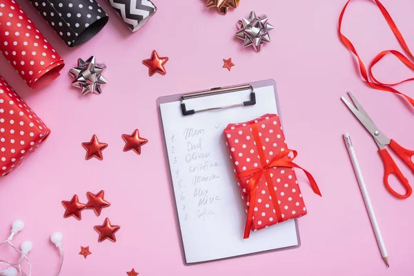 List of names and wrapping Christmas gifts flat lay on pink background. Top view of Christmas prepare concept.