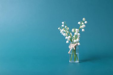 Lilies of the valley bouquet in a mini glass jar on a turquoise background with copy space. Minimalistic spring still life.
