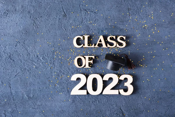 Class of 2023 concept. Wooden number 2023 with graduated cap on dark concrete background with tinsel.