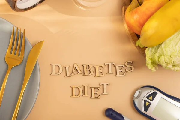 Diabetes diet text with food plate and cutlery, glucose meter on beige background flat lay, top view.