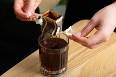 Kadın elleri cam bir fincanda demlenmiş bir kahve torbası tutuyor..