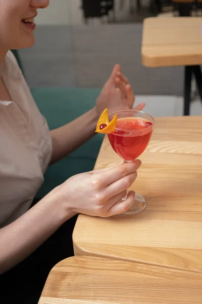 Alcoholic cocktail in stemmed glass in female hand on wooden cafeteria table. Drinking cocktail concept