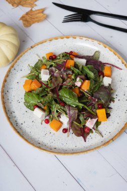 Yeşillikli salata, balkabağı, yumuşak peynir ve ahşap arka planda çatal bıçak takımı.