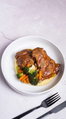 Kırmızı soslu bir tabak biftek, sebzeli ve patates püresi ve siyah çatal bıçak..