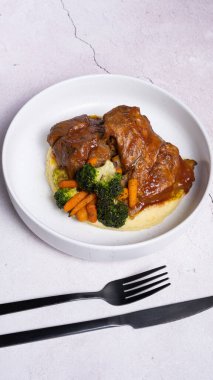 Kırmızı soslu bir tabak biftek, sebzeli ve patates püresi ve siyah çatal bıçak..