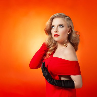 Şık sarı saçlı, canlı kırmızı elbisesi ve kalın makyajı olan çekici bir genç kadın. Canlı turuncu bir arka plana karşı zarif bir şekilde poz veriyor, sofistike ve özgüven yayıyor..