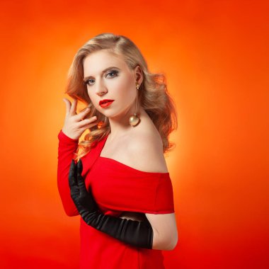 Şık sarı saçlı, canlı kırmızı elbisesi ve kalın makyajı olan çekici bir genç kadın. Canlı turuncu bir arka plana karşı zarif bir şekilde poz veriyor, sofistike ve özgüven yayıyor..