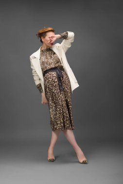 Leopar desenli elbisesi ve topuklu ayakkabısı olan zarif olgun bir kadın stüdyoda poz veriyor. Özgüven ve zarafetle şık moda portresi.