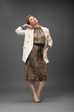 Leopar desenli elbisesi ve topuklu ayakkabısı olan zarif olgun bir kadın stüdyoda poz veriyor. Özgüven ve zarafetle şık moda portresi.