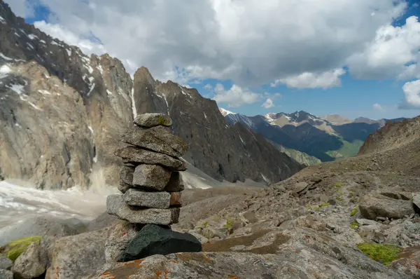 Stacked rock cairn stands prominently against a backdrop of towering snow-dusted peaks, under a vast blue sky with scattered clouds, overlooking a valley with hints of green foliage.