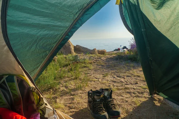 View of the coastal landscape from inside the tent with sleeping bags and a backpack. Hiking boots. Sea merges with a blue sky. To the right of the tent, stones and other camping items are arranged