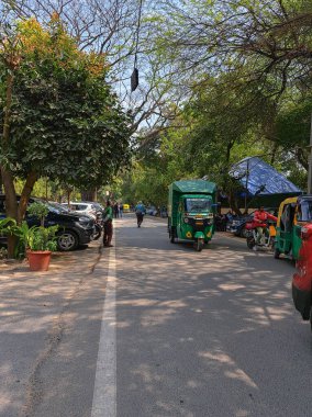 25 Nisan 2024, Yeni Delhi, Hindistan. Sokak hayatı, yayalar, park edilmiş arabalar. Moto-rickcart 'lar yolcu taşıyor. Yoğun bir trafik. Yemyeşil ağaçların yolu, kentsel atmosfer