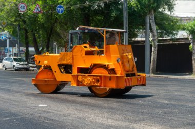Parlak turuncu sıkıştırma makinesi, asfalt silindirler şehir yolunda yeni asfalt kaldırımlar, trafik işaretleriyle çevrili, park edilmiş araçlar. Etkin yol geliştirme ve bakım çalışmaları.