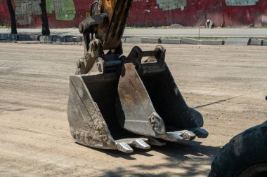 Tozlu caddeye yerleştirilmiş ağır kazıcı kovası, şehir ortamında asfalt döşeme hazırlığı kapsamında not verme, kazma ve malzeme kaldırma ile meşgul.