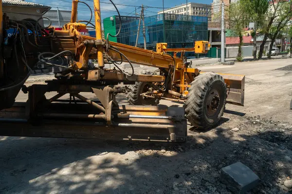 Şehir caddesinde çalışan büyük çaplı makineler, asfalt uygulamasından önce çakıl zemini düzleştiriyor ve düzleştiriyor, binalar, inşaat malzemeleri, elektrik hatları ve temiz gökyüzü arkaplanı ile çevrili..